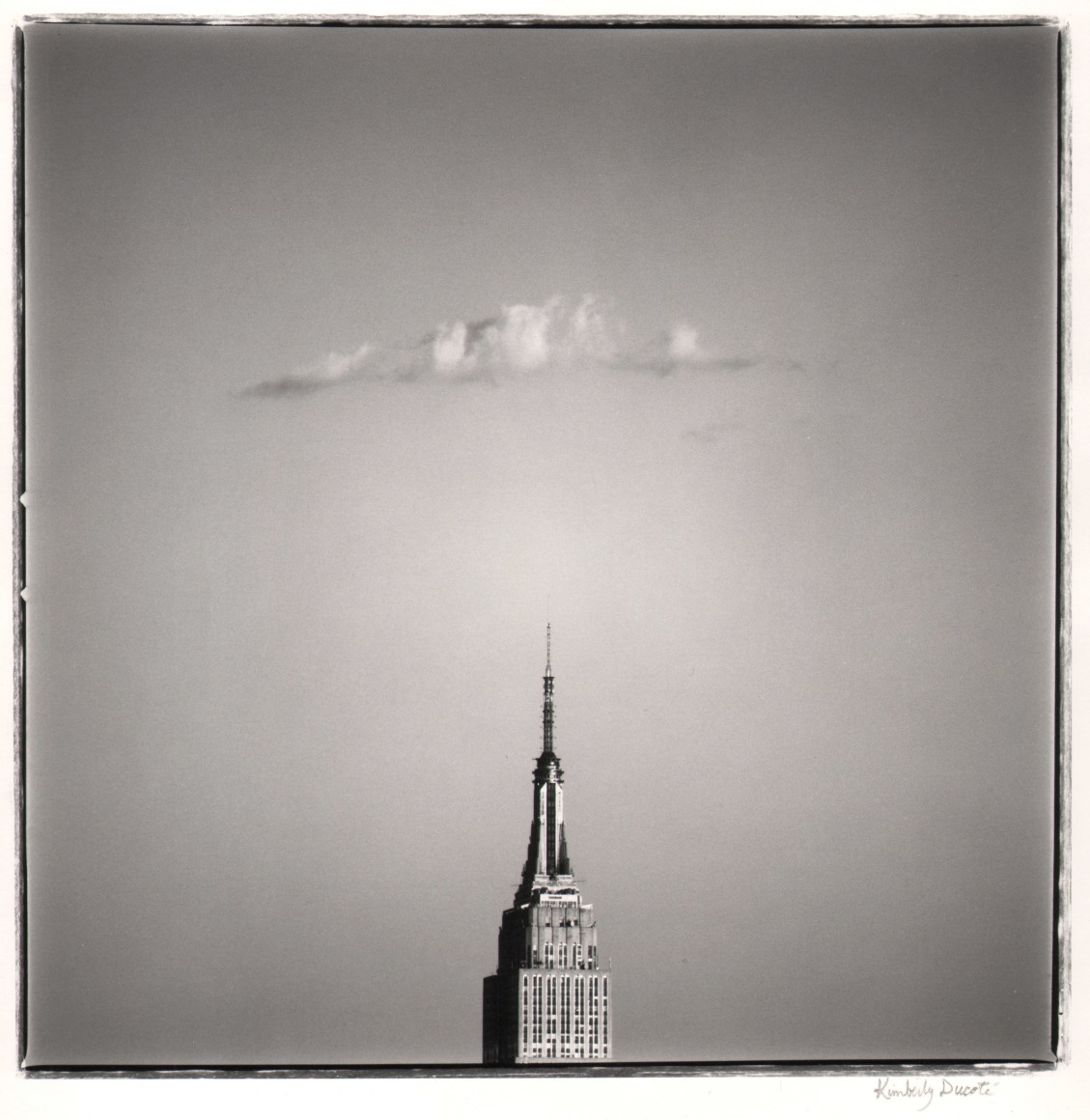 07. Kimberly Ducot&eacute;, Empire State Building, ​1990. Distant, head-on view of the top of the Empire State Building, which occupies the lower center of the frame. Above it is a single cloud.