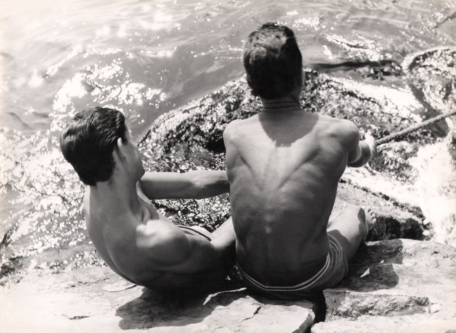 Mario Cattaneo, Lo Sforzo, ​c. 1960. Two shirtless young men, backs to the camera, sit on a rock by the water, pulling a rope that extends out of the right of the frame.