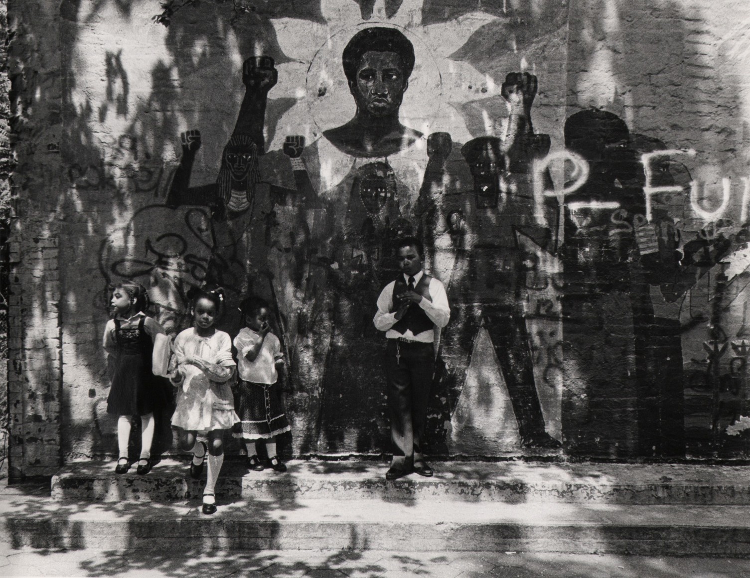 24. Beuford Smith, Kids in Park, NYC, ​c. 1970. Three young girls and one boy lean against a wall with a mural and graffiti in speckled light.