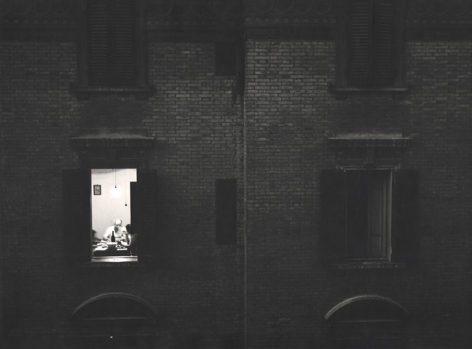 Nino Migliori, Gente dell'Emilia, 1953. Exterior view of an apartment building. One window is lit and a family is seated at the dinner table.