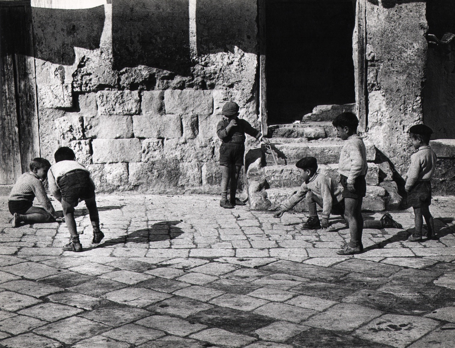 Giuseppe Bruno, Viaggio del Sud, Matera, ​1958. Six young boys play on the cobbled street in front of the doorway of a stone house. Three are standing; three crouch low to the ground.