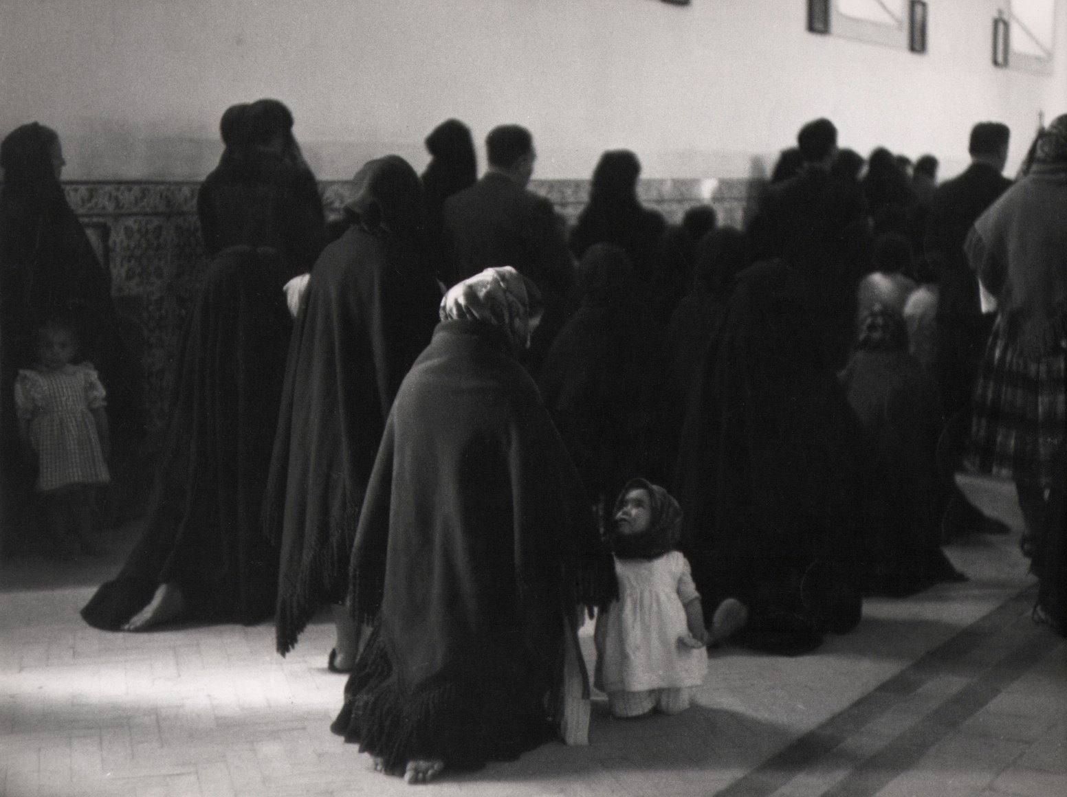43. Sabine Weiss, La Messe, Portugal, 1954. A crowd of dark-cloaked figures in a hall; a young girl kneels with one cloaked woman in the center of the frame.