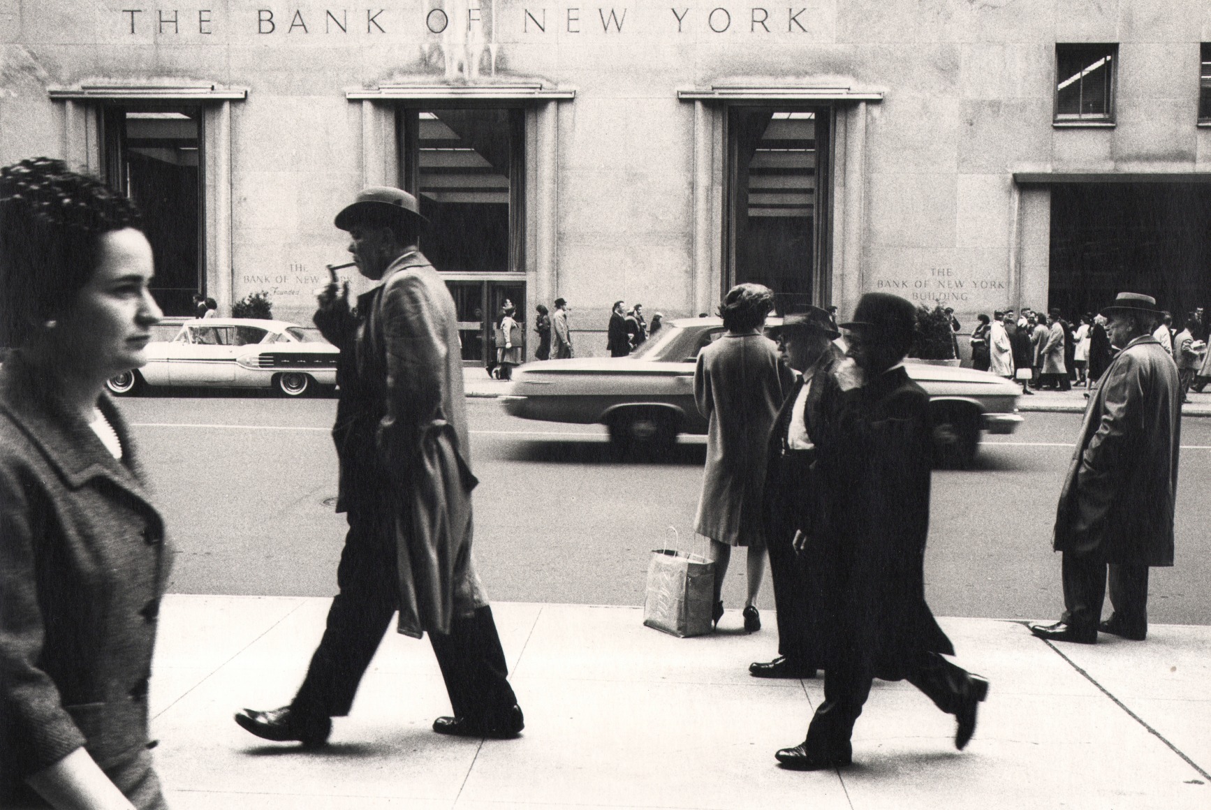 05. Simpson Kalisher, Untitled, ​c. 1959. Pedestrians walk horizontally across the frame on a sidewalk across the street from the Bank of New York, seen in the background.