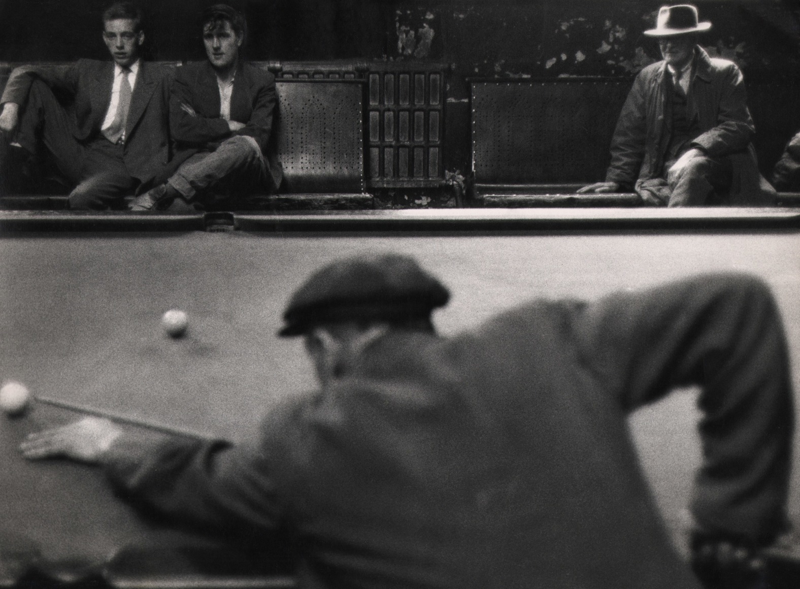 29. John Bulmer, The billiards room at Nelson, Lancashire, ​c. 1966. A man in the foreground is out of focus, back to the camera, leaning over the pool table. Three men watch on from benches in the background.