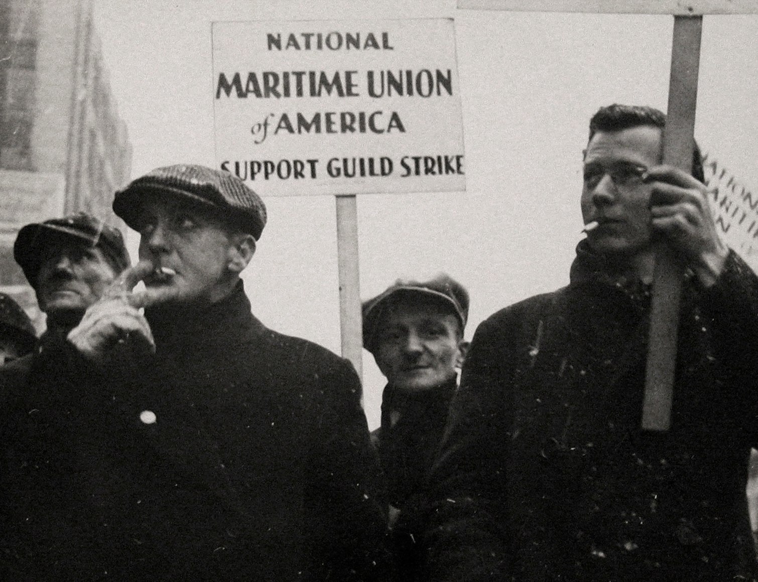 19. Gordon Coster, W.P.A. Parade, Chicago, 1939. Four men walk in black coats on a snowy day, smoking cigarettes. One holds a sign that reads &quot;National Maritime Union of America Support Guild Strike&quot;