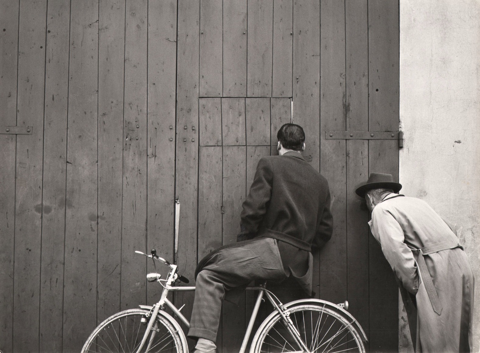 Stanislao Farri, Curiosit&agrave;, ​1958. One man on a bicycle and one standing peer into the spaces between the wooden slats of a door.