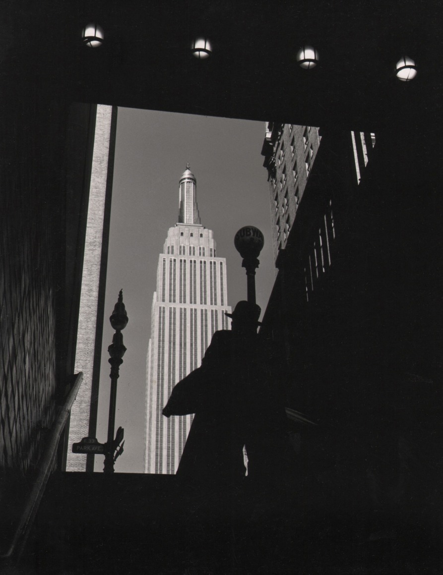 19. John C. Hatlem, Empire State Building, ​c. 1935. View from the bottom of subway entrance stairs, looking out and up towards the Empire State Building. A figure in a hat stands on the stairs and is silhouetted against the building.