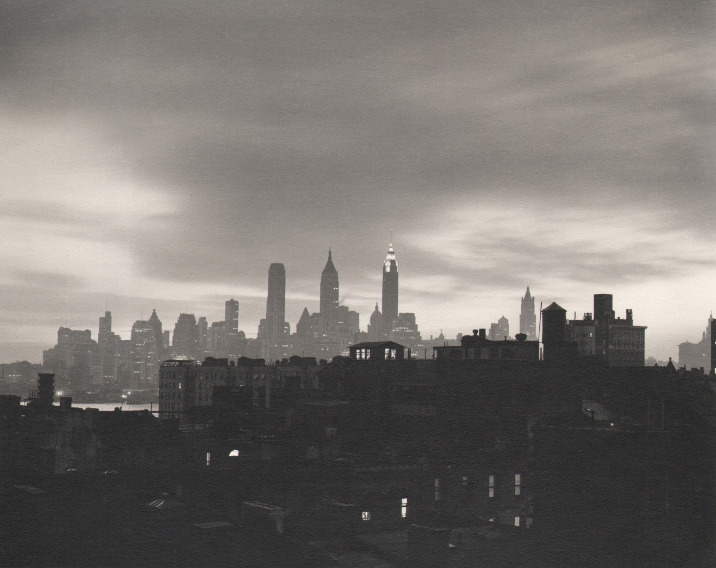 35. John C. Hatlem, New York City Skyline, ​c. 1935. Dark, mostly silhouetted view of the city from across a river. Dark buildings in the foreground. The Empire State is in the center background against a cloudy sky.