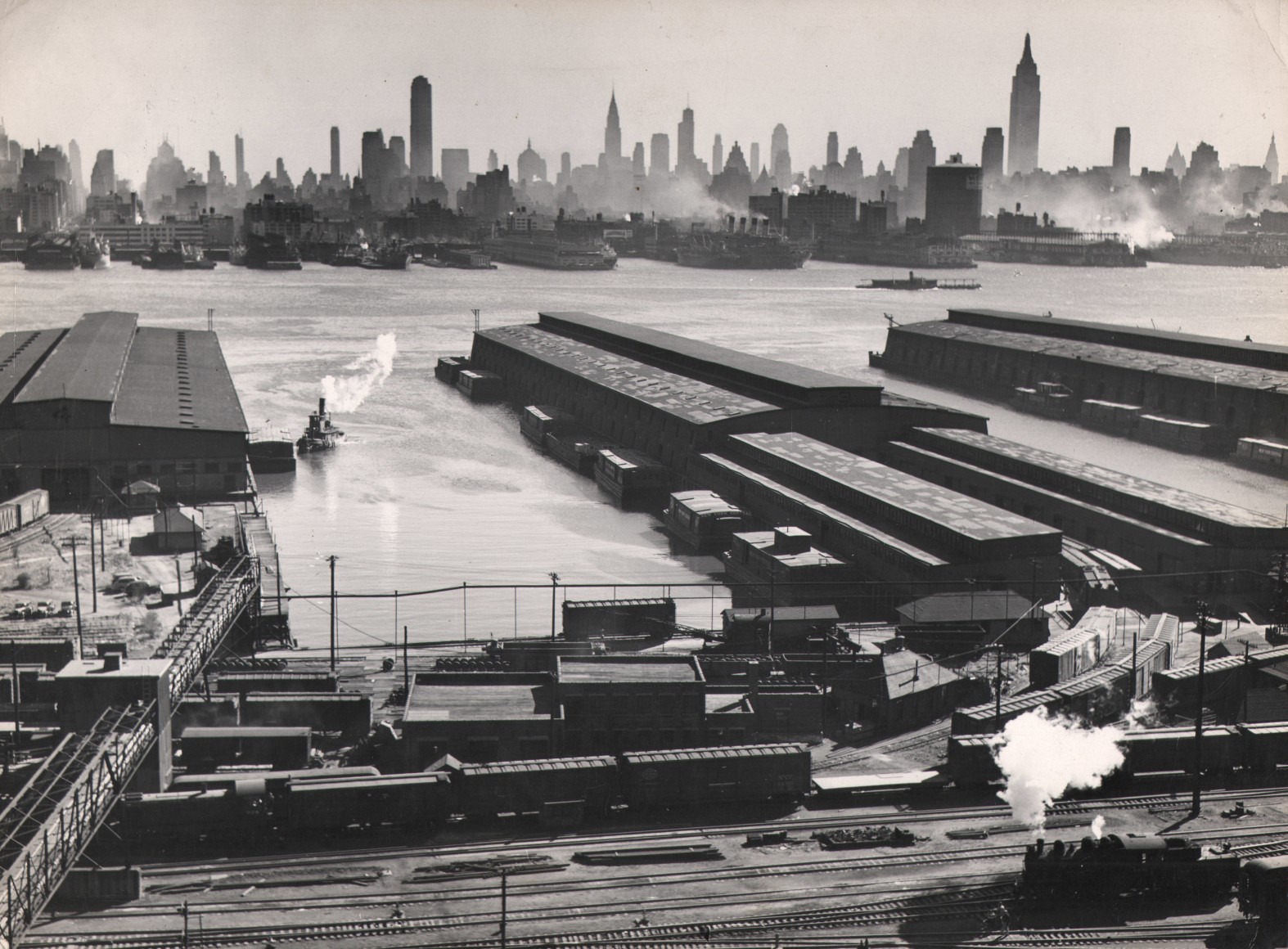 6B. Esther Bubley, Untitled, 1946. Train tracks in the foreground. Piers extend into the a river in the midground with the city skyline in the background.