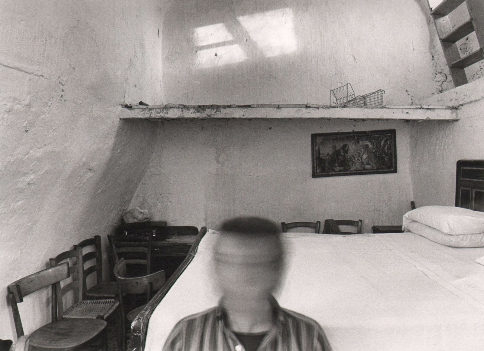 Mario Cresci, Untitled, ​1979. A figure's head, blurred with motion, and shoulders are in the center foreground of a small bedroom with a bed against the right wall and chairs along the left and rear walls.