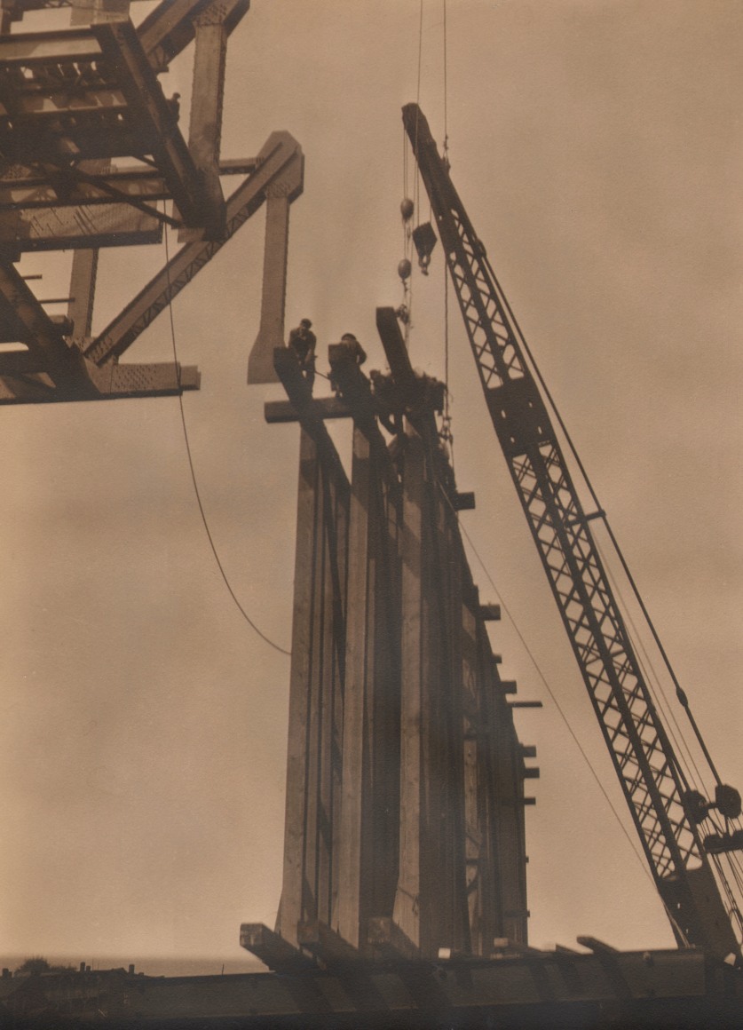 19. Margaret Bourke-White, Untitled, c. 1930. Men working atop a series of beams, photographed from below. A crane rises up along the right side of the frame.