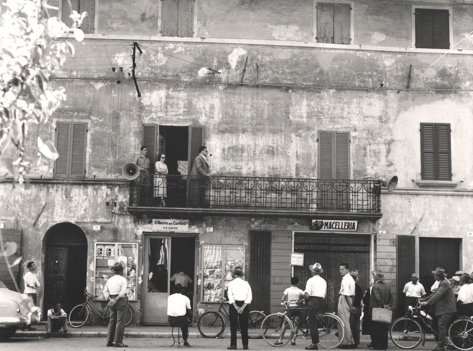 Nino Migliori, Meeting in the Province, 1957. People gather in the street as a man speaks into a microphone from a balcony.