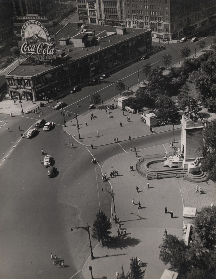 30. Charlotte Brooks, Columbus Circle, 1943. Aerial view of the street and sidewalk; a fountain with statues on the right of the frame. A large Coca-Cola sign is on a building at the top left of the frame.