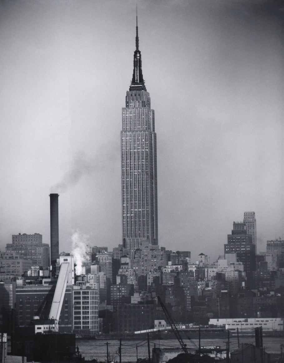 25. Allen Locker, Empire State Building, ​c. 1965. The Empire State Building fills the center of the frame against a cloudy sky in a skyline view from across a river.