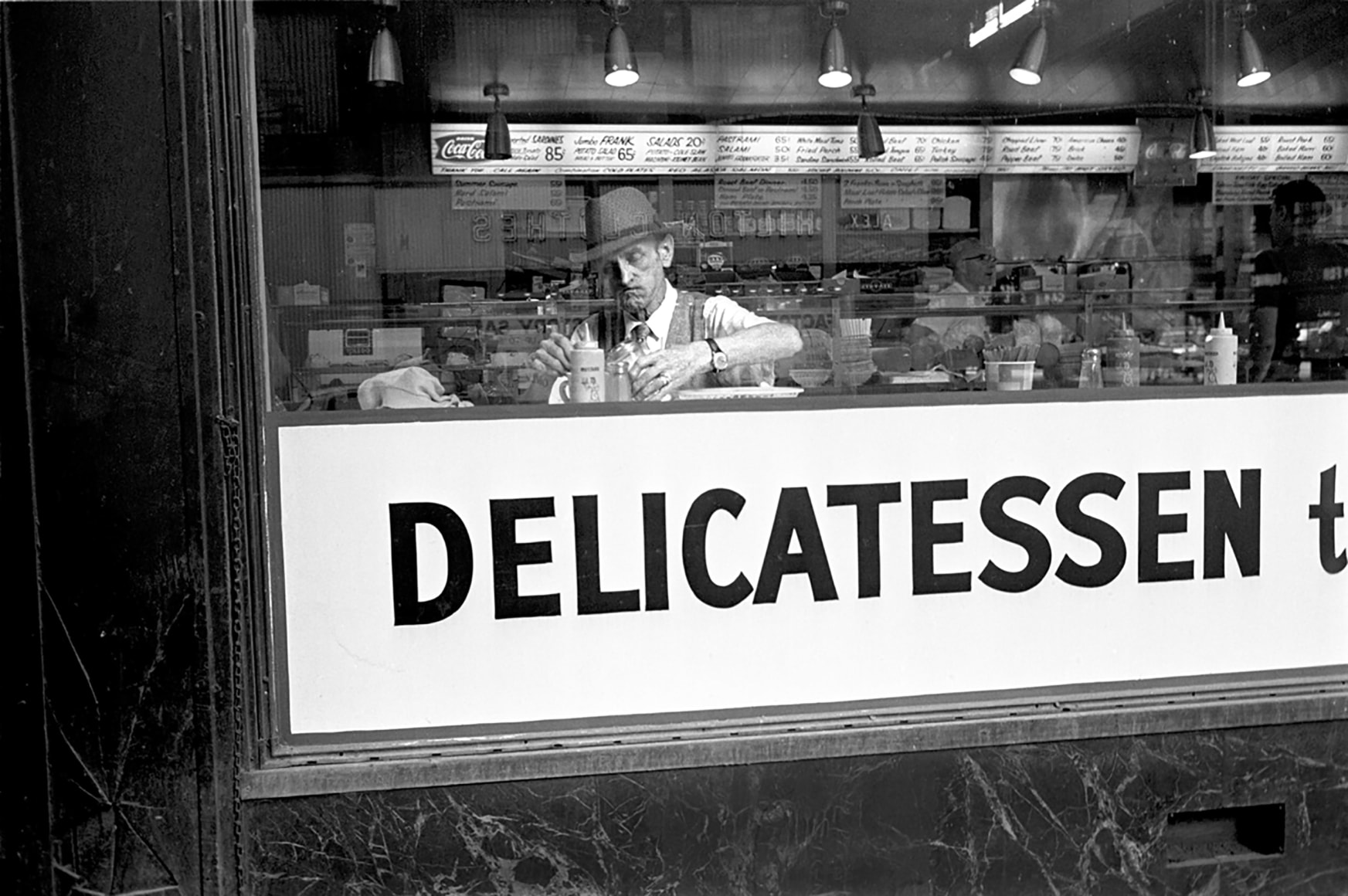 11. Simpson Kalisher, Untitled, ​c. 1960. Man seated at deli counter behind window that reads &quot;Delicatessen&quot;