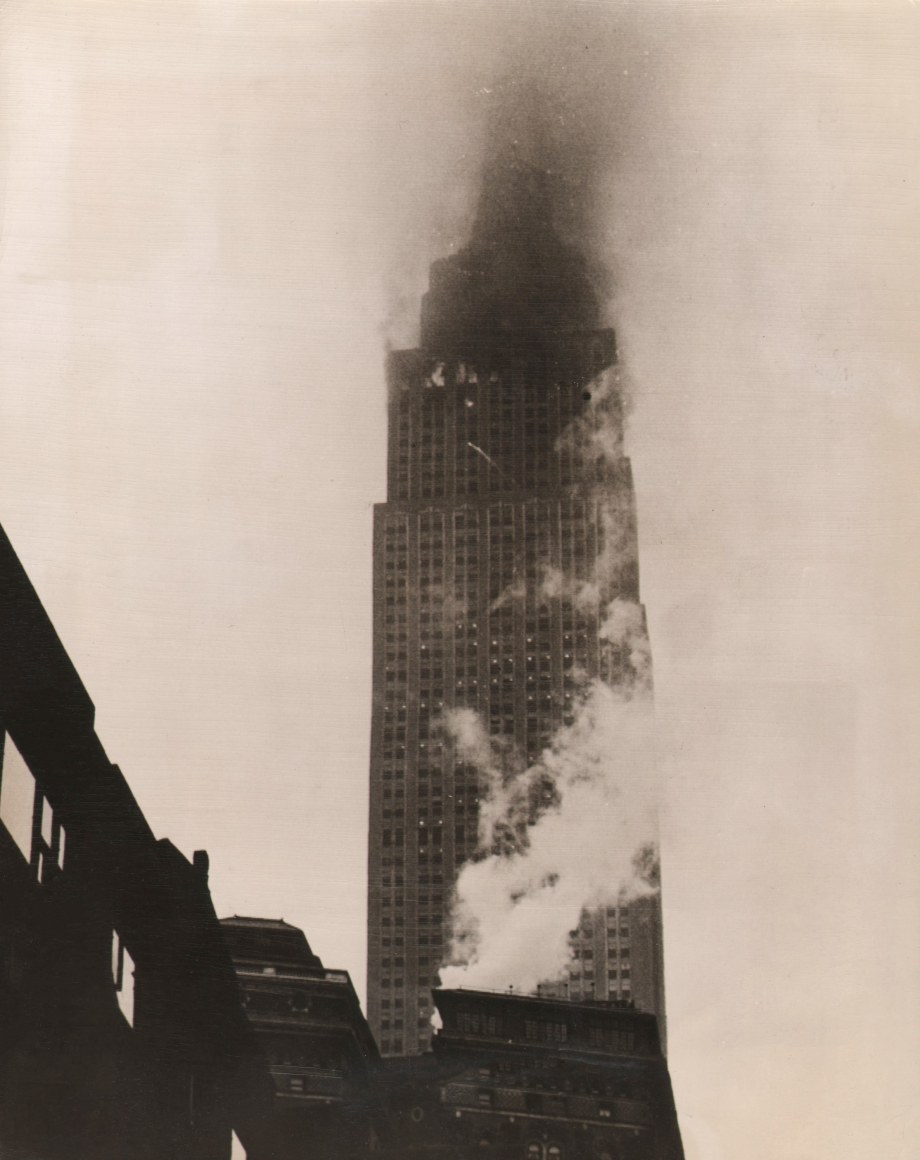 02. Anonymous, Empire State Afire After Plane Crash, ​1945. Street view of a skyscraper with smoke emitting from the upper floors.