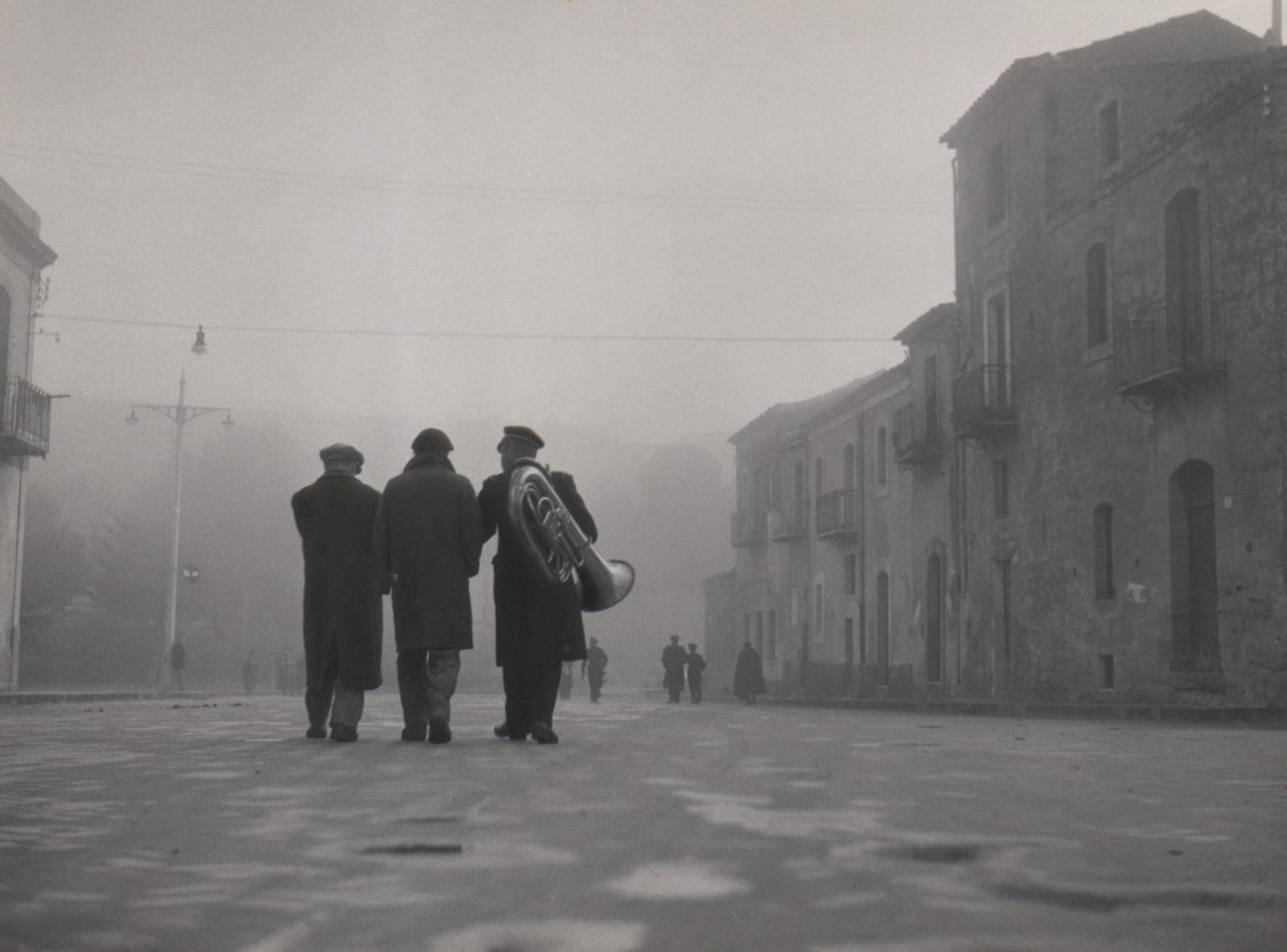 Fosco Maraini, Grey Sunday, ​1952. Three men in black walk away from the camera on a hazy street; one carries a tuba on his back.