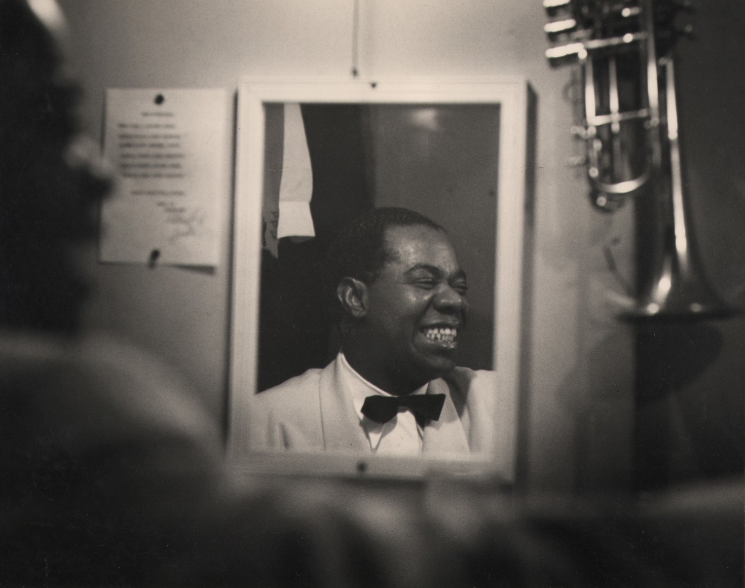 Bob Willoughby, Louis Armstrong, ​1950. Subject is seen smiling in a mirror reflection, head turned to the right.