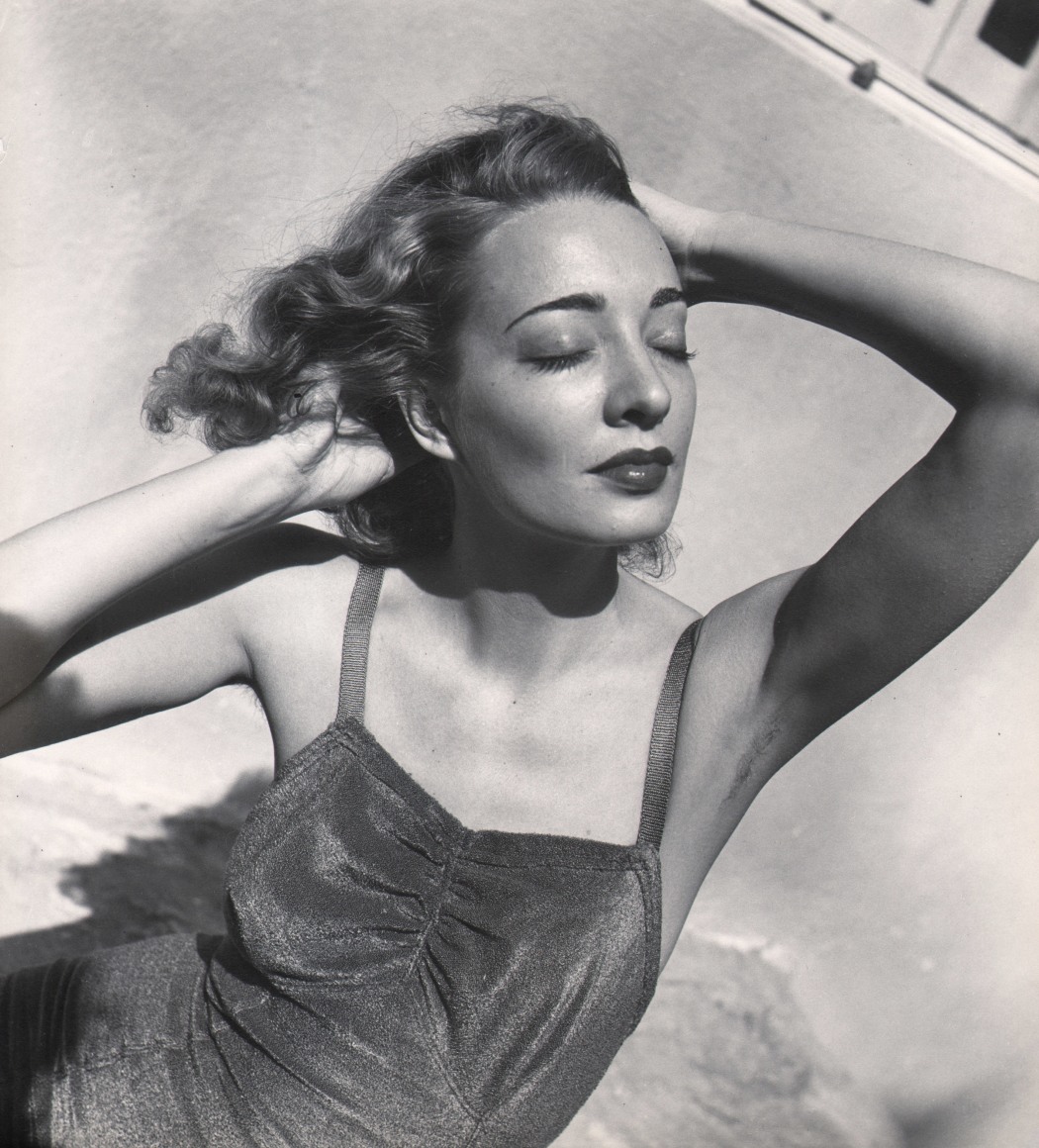 23. Louise Dahl-Wolfe, Untitled, c. 1940. A woman in a swimsuit with both hands in her hair. Her face is tilted towards the upper right of the frame with eyes closed.