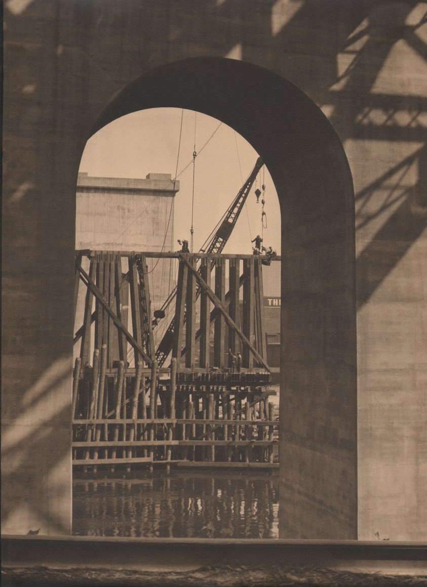 17. Margaret Bourke-White, Untitled, c. 1929. Looking through the arch of a bridge to a crane and beams rising out of the water.