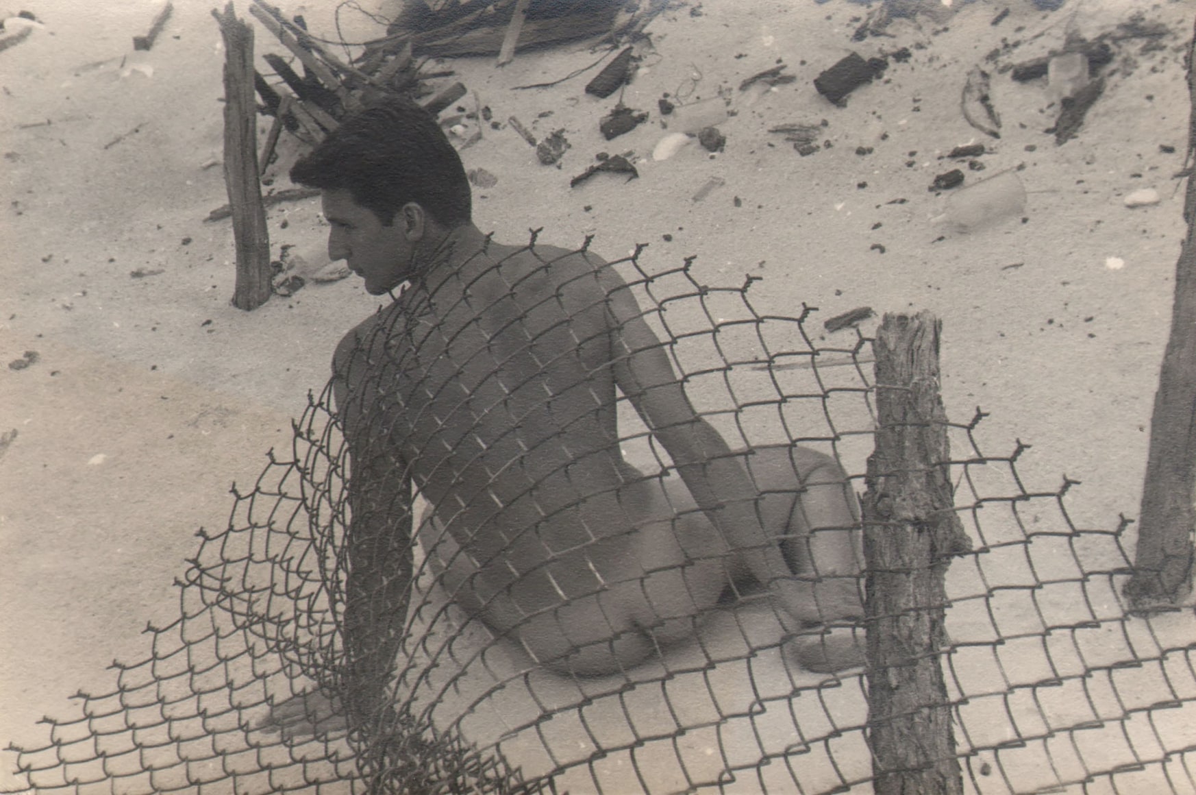 PaJaMa, Jack Fontan, ​c. 1945. Male nude seated in the sand, back to the camera, behind a chain link fence, head turned left.
