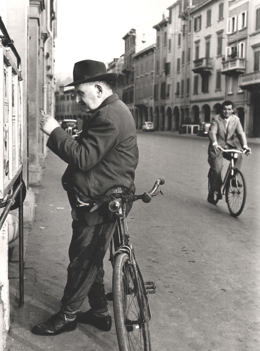 Nino Migliori, People of Emilia, 1950. Street scene featuring a man leaning on his bicycle in the foreground while another man rides a bicycle in the background.
