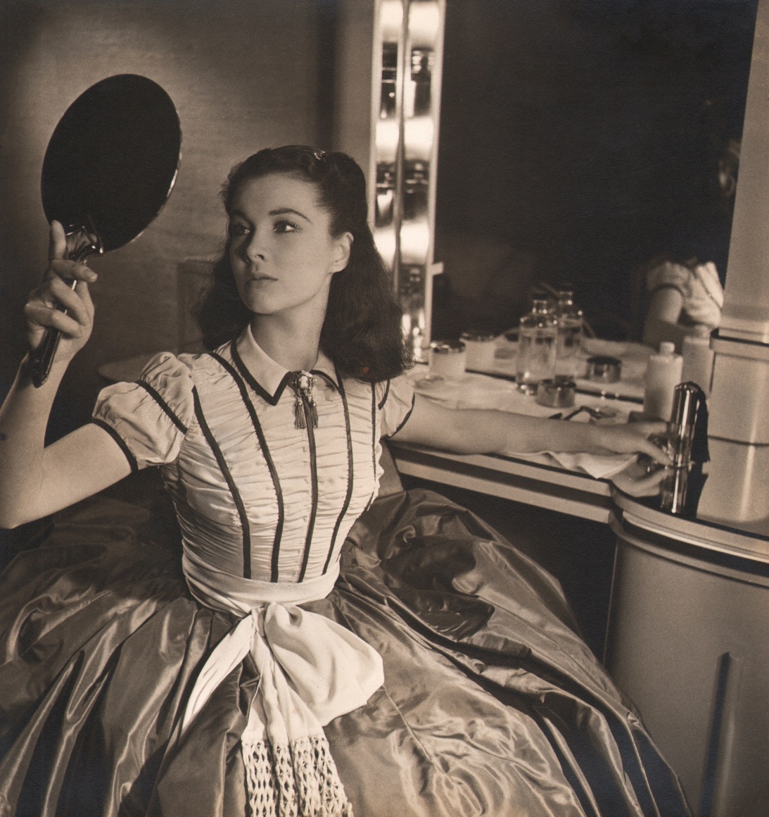 22. Louise Dahl-Wolfe, Vivien Leigh, c. 1939. A woman seated at a vanity holds up a small mirror to the left of the frame to look at her reflection.