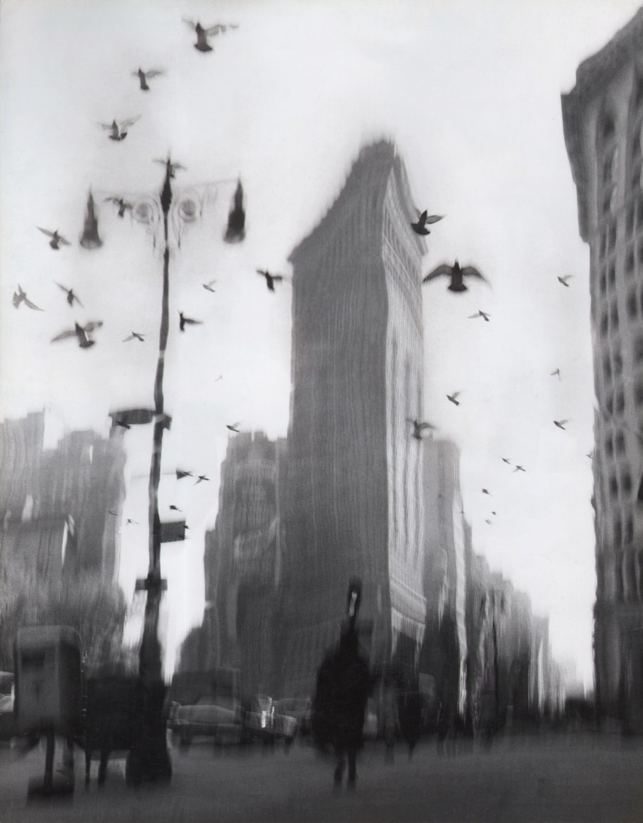 32. David Attie, Flatiron Building, ​c. 1955. Blurry view of figures in the street and birds in flight in front of the Flatiron Building in the center midground.