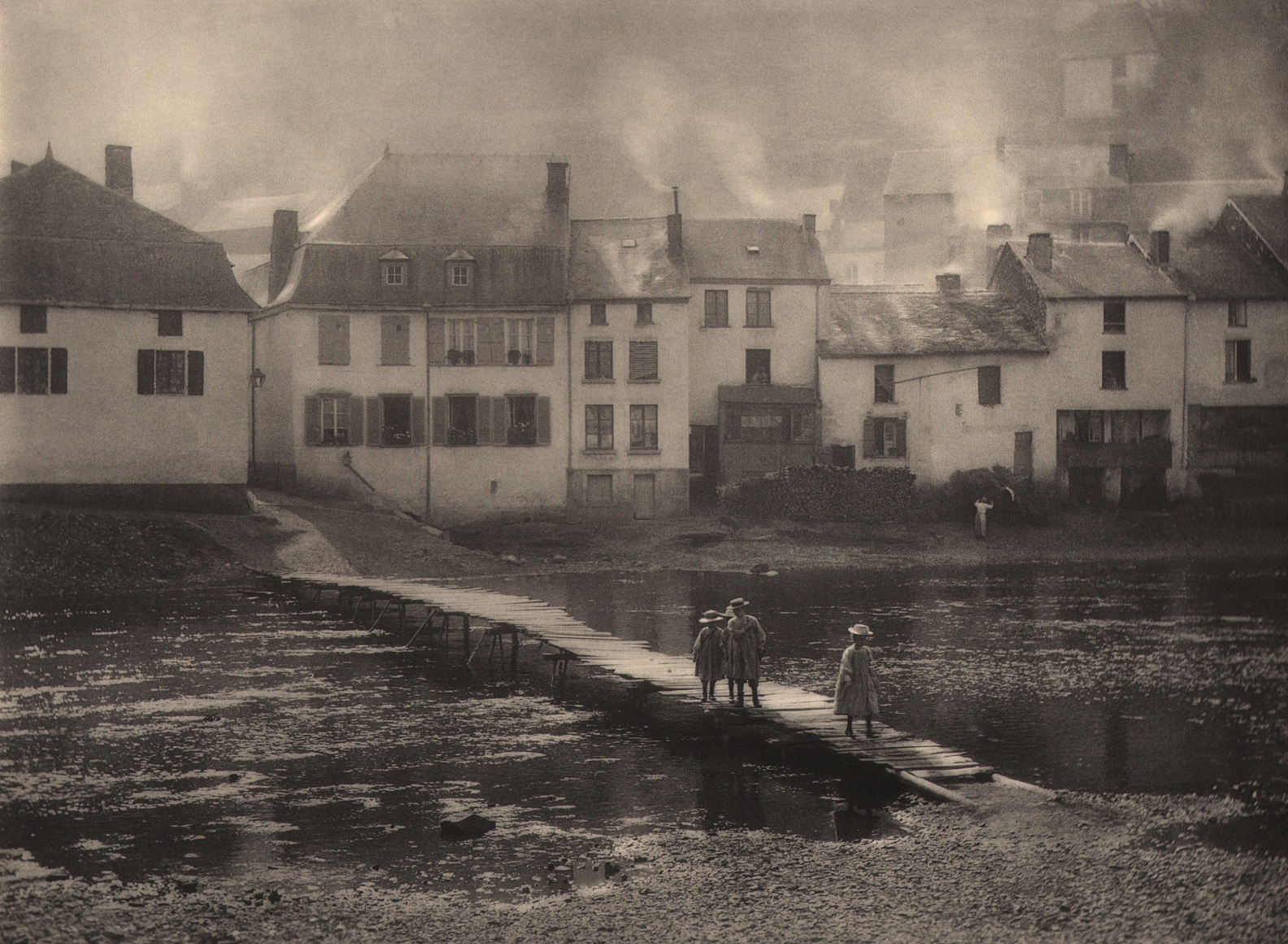 15. L&eacute;onard Misonne, On alume le jeux, 1924. Three young girls walk across a small body of water on a wooden dock. Waterside homes with smoke rising above them in the background. Sepia-toned print.