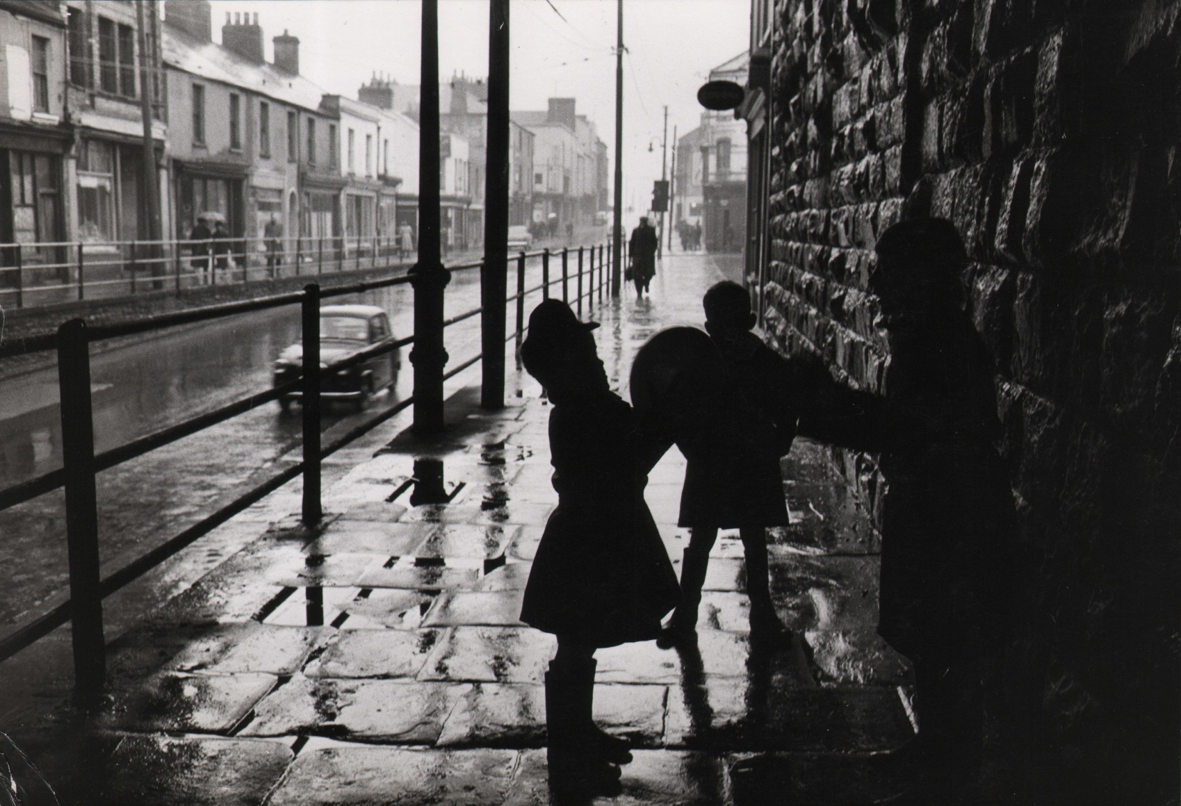 16. Patrick Ward, Street in the Tiger Bay dockland area of Cardiff, Wales, c. 1961&ndash;1966. Silhouettes of three children in the foreground right on a wet cobbled sidewalk.