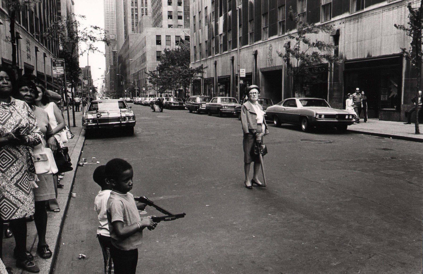 17. Anthony Barboza, NYC, ​1970s. Street scene. Two small black children holding toy guns in the foreground with a line of women on the sidewalk to the left. A middle-aged white woman stands in the center of the car-lined street.