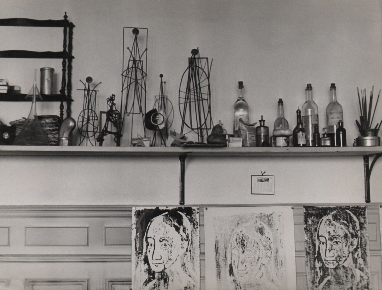 Brassa&iuml;, Atelier de Picasso, ​c. 1933. A shelf on a wall featuring various glass bottles, wire sculptures, and paint brushes. Three prints of the same face hang below it.