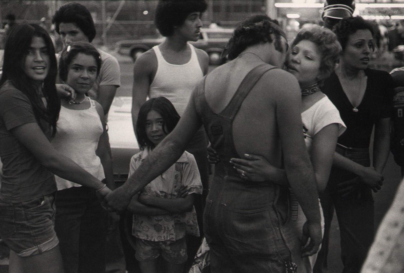 12. Anthony Barboza, Coney Island, NY, 1970s. Crowd of people. Central figure is a man in overalls with back to the camera. He holds one woman's hand while embracing another.