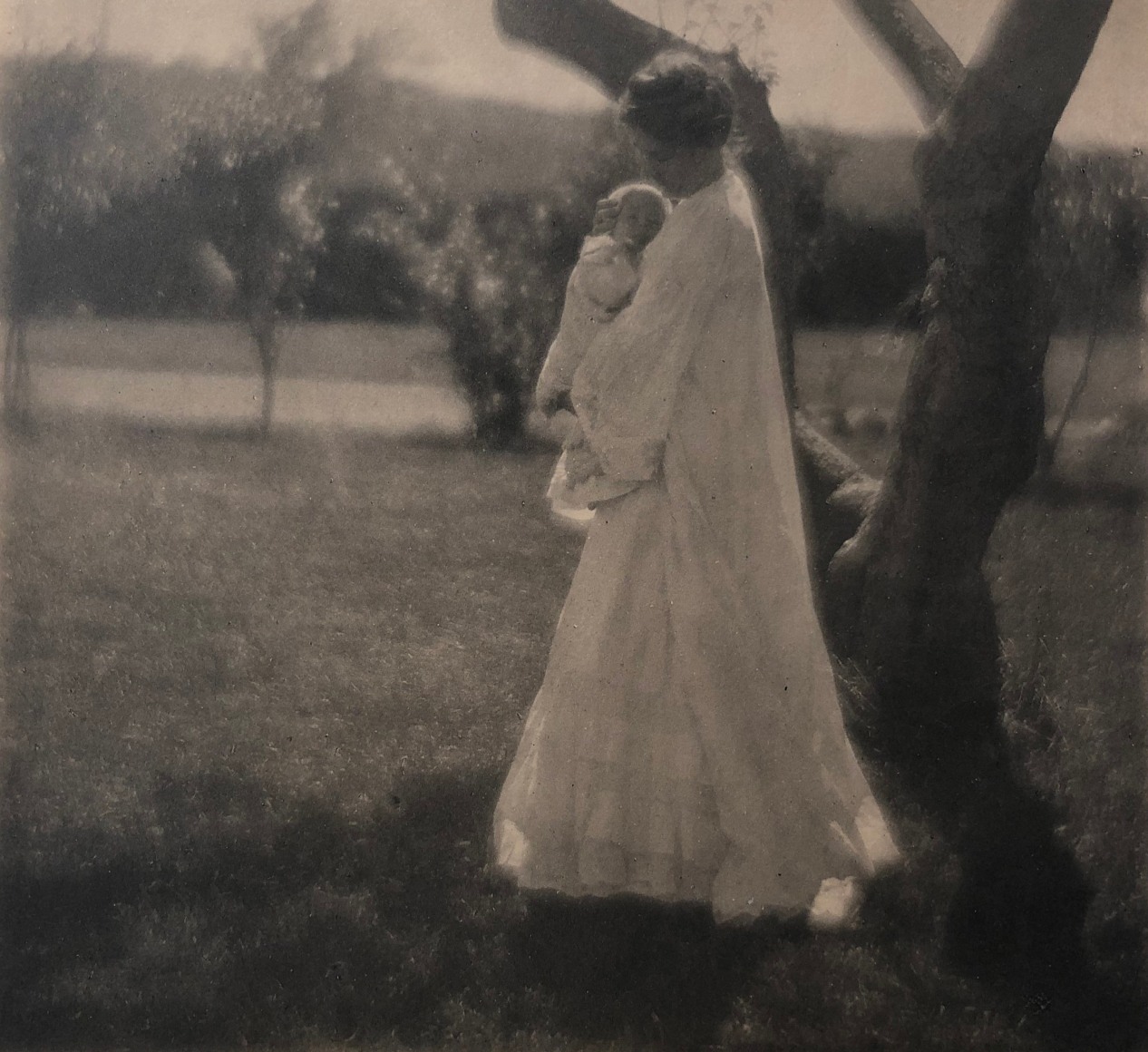 04. Gertrude K&auml;sebier, Blossom Day, ​1904. A woman in a long white dress stands beneath a tree holding a baby to her chest.