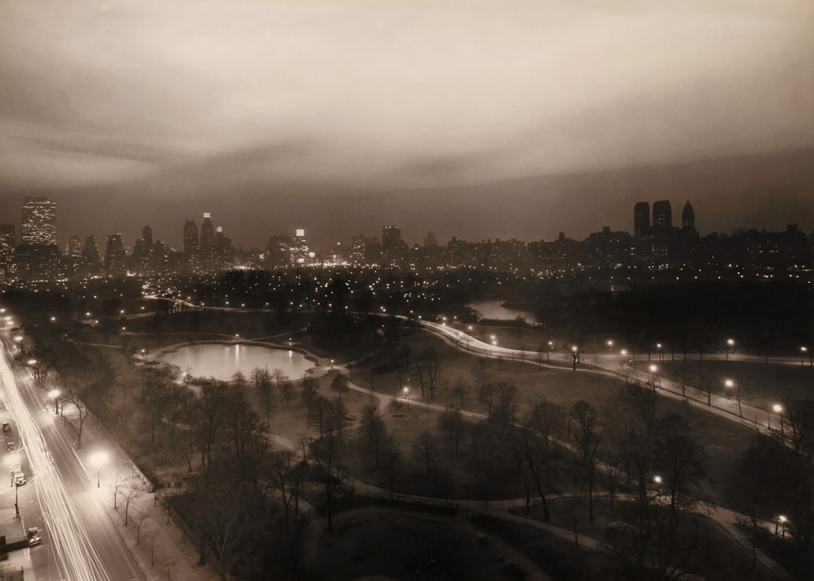 Paul J. Woolf, Central Park Looking South, ​c. 1936. Night time cityscape with the park filling the lower half of the frame, a street running up the lower left. Tall buildings across the center of the frame and an overcast sky above.