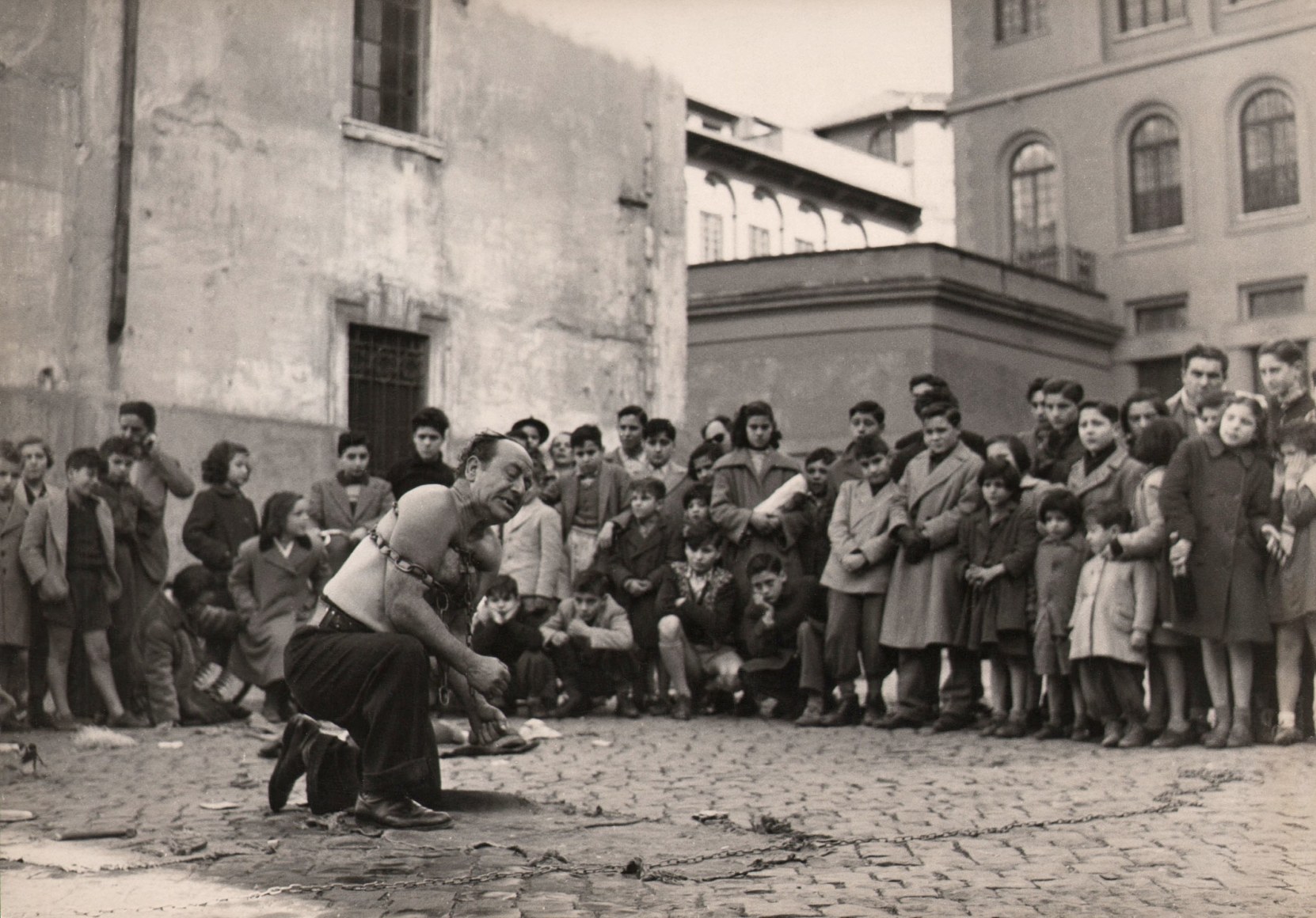 Franco Pinna, Roma - Panico, ​c. 1955. A street performer kneels with a thick metal chain around his chest and arms, a large group of children looking on in the background.