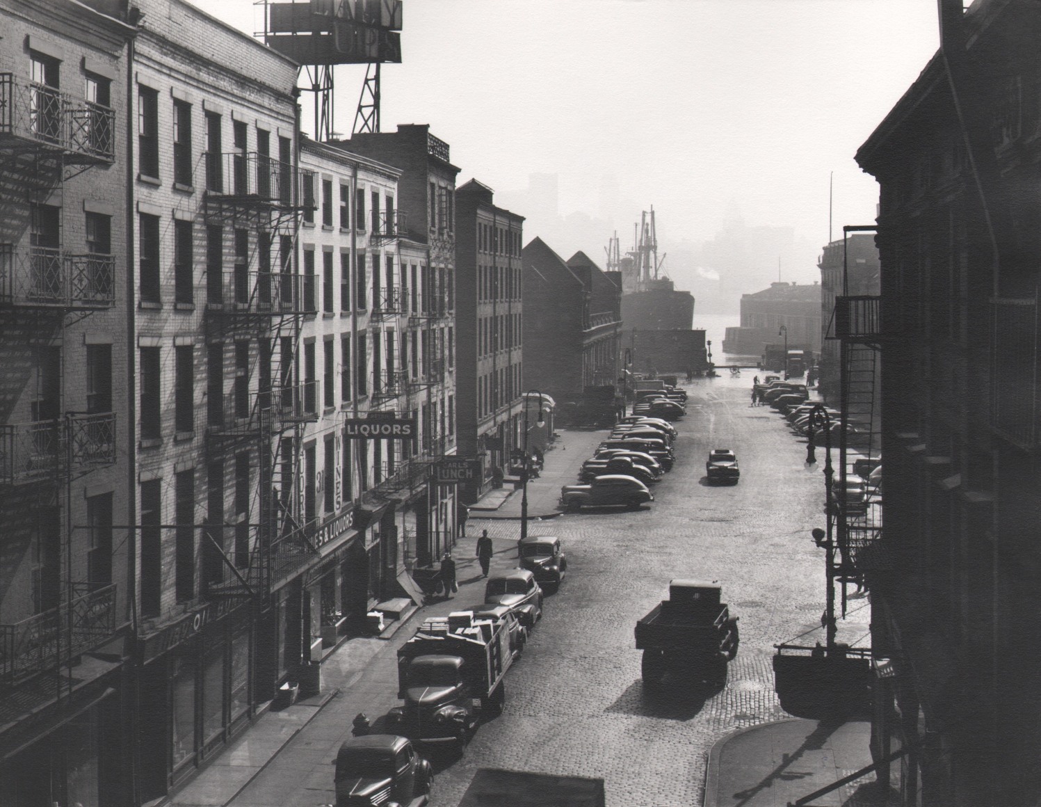 46. Esther Bubley, New York Harbor: View looking East on Fulton Street from Third Avenue El platform, ​1946. Elevated view of a car-lined street ending in water and boats.