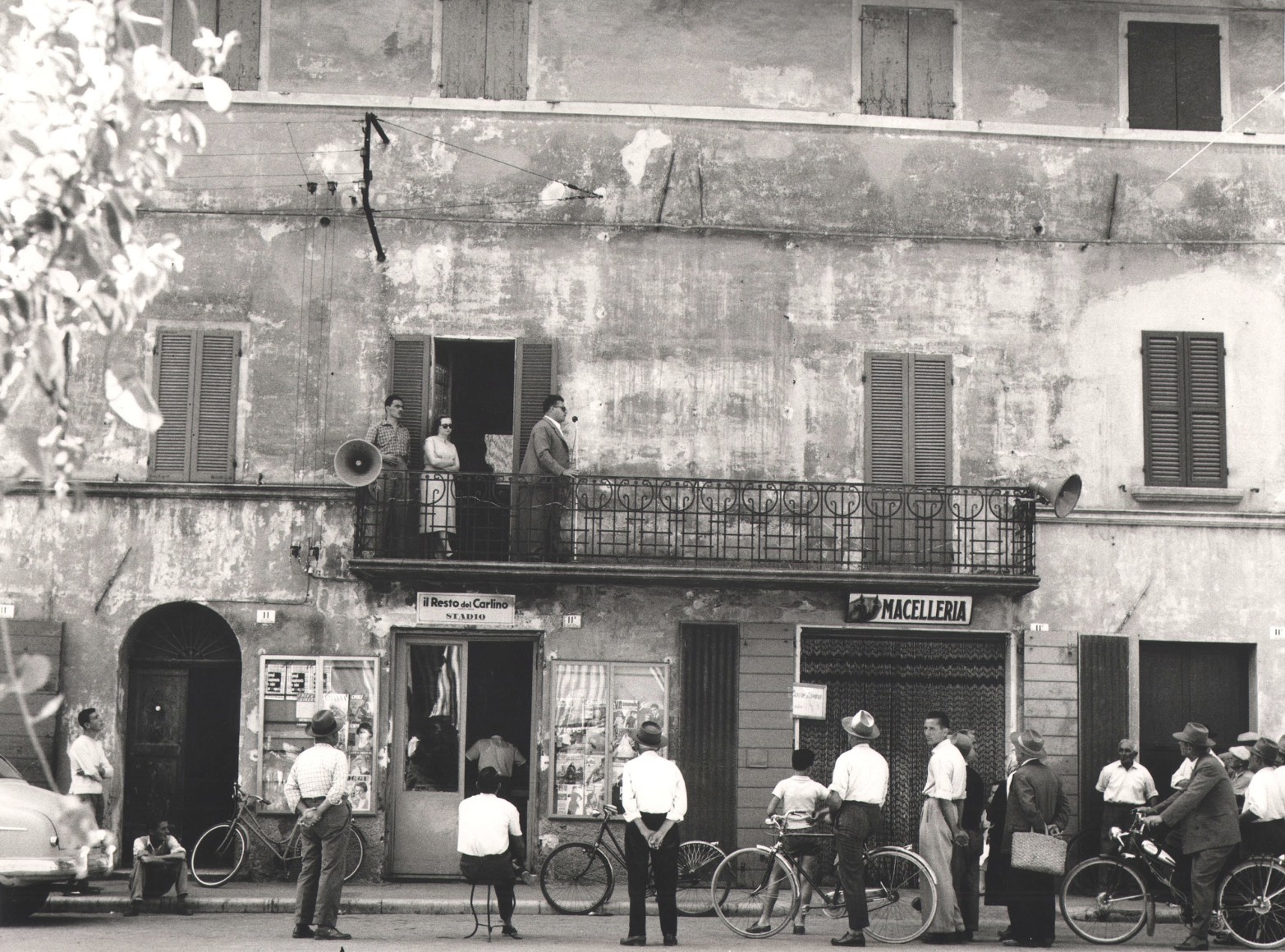 Nino Migliori, Gente dell'Emilia, 1957. People gather in the street as a man speaks into a microphone from a balcony.