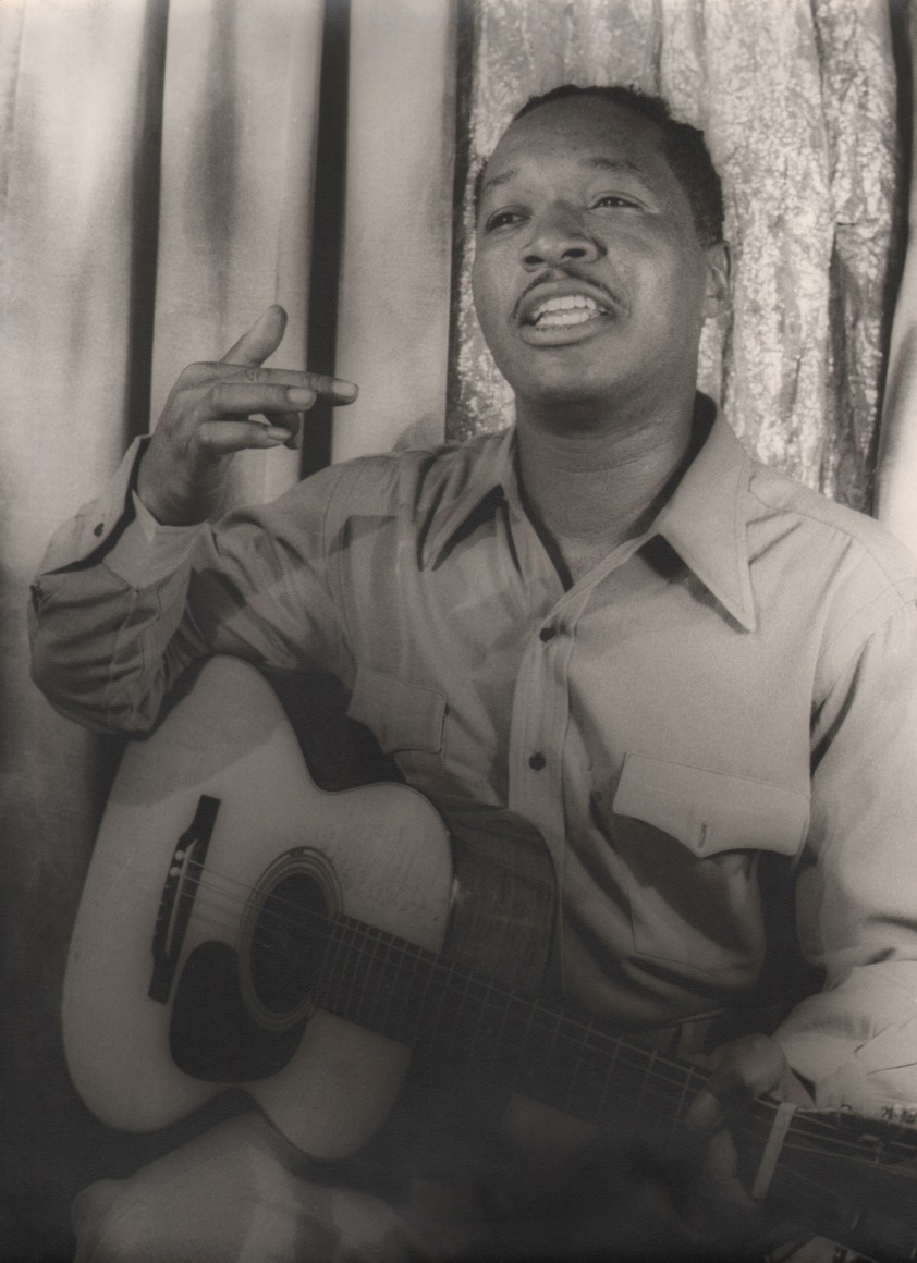 30. Carl Van Vechten, Josh White, ​1946. Seated portrait with subject holding a guitar in his lap with on hand on the neck and the other raised in front of him.