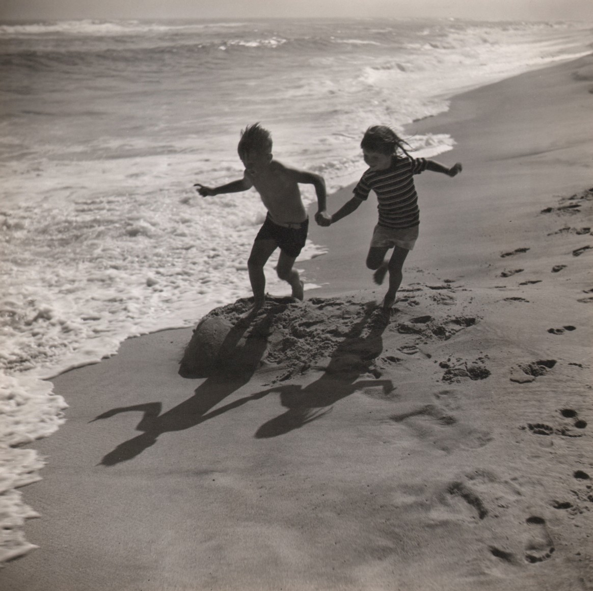 45. Toni Frissell, Untitled, c. 1947. Two children holding hands and running on the beach. The frame is divided from lower left to upper right by the sea meeting the sand.