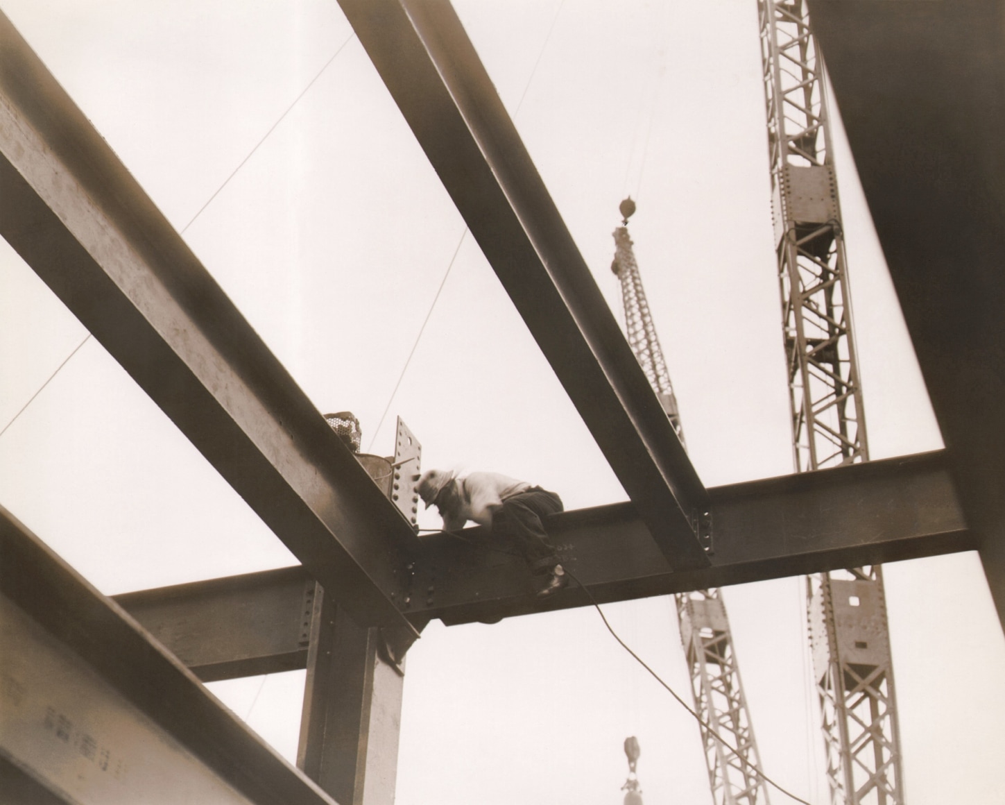 Paul J. Woolf, Rockefeller Center Construction, ​c. 1933. Detail of structural metal supports and a construction worker seated on one of the horizontal pieces.