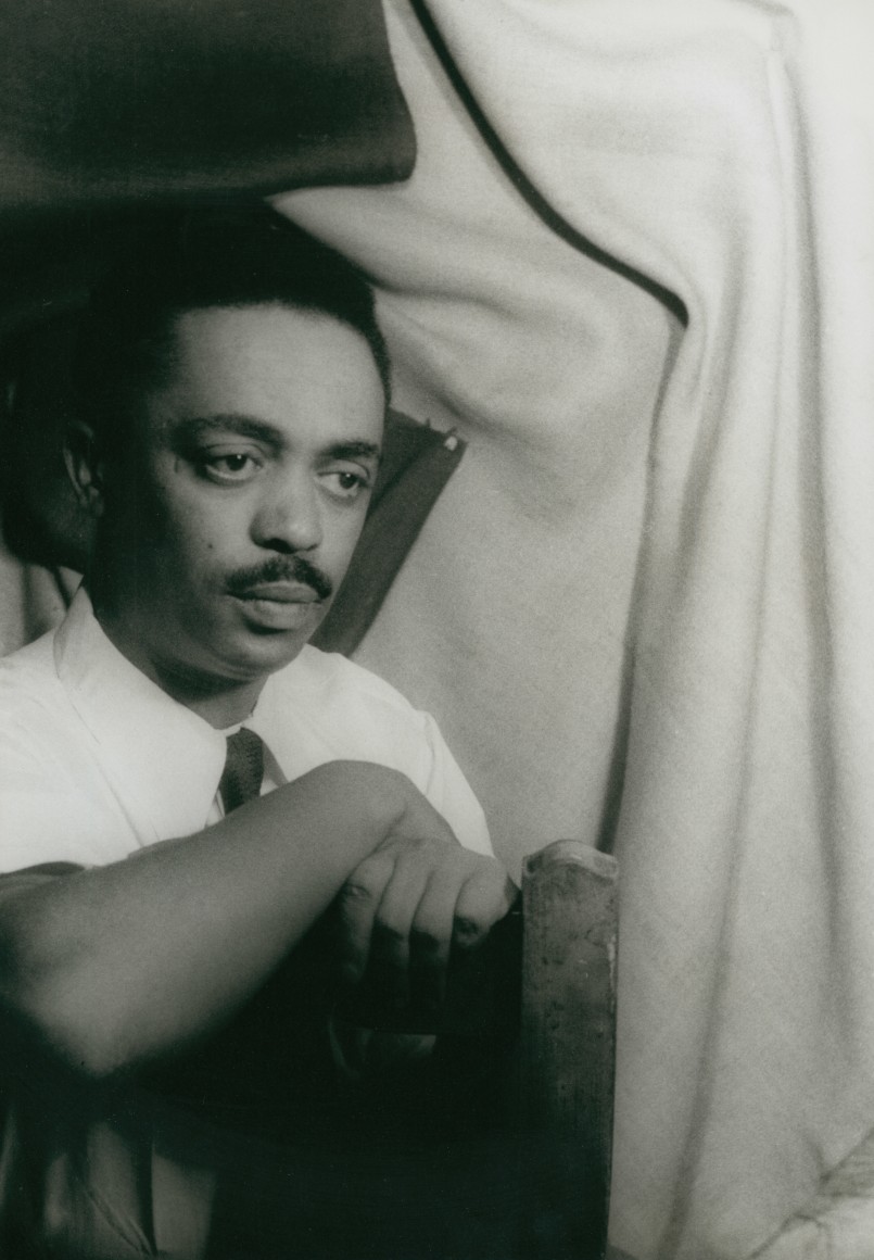 23. Carl Van Vechten, Peter Abrahams, ​1955. Seated portrait with subject at 45 degrees towards the left of the frame, arms crossed over the back of a wooden chair.