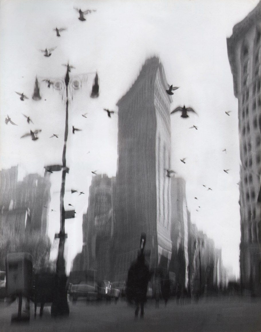 26. David Attie, Flatiron Building, ​c. 1958. Blurred/distorted photo of a triangular building against a gray sky. Various figures and cars can be seen in the lower third of the print, below a number of birds taking flight.