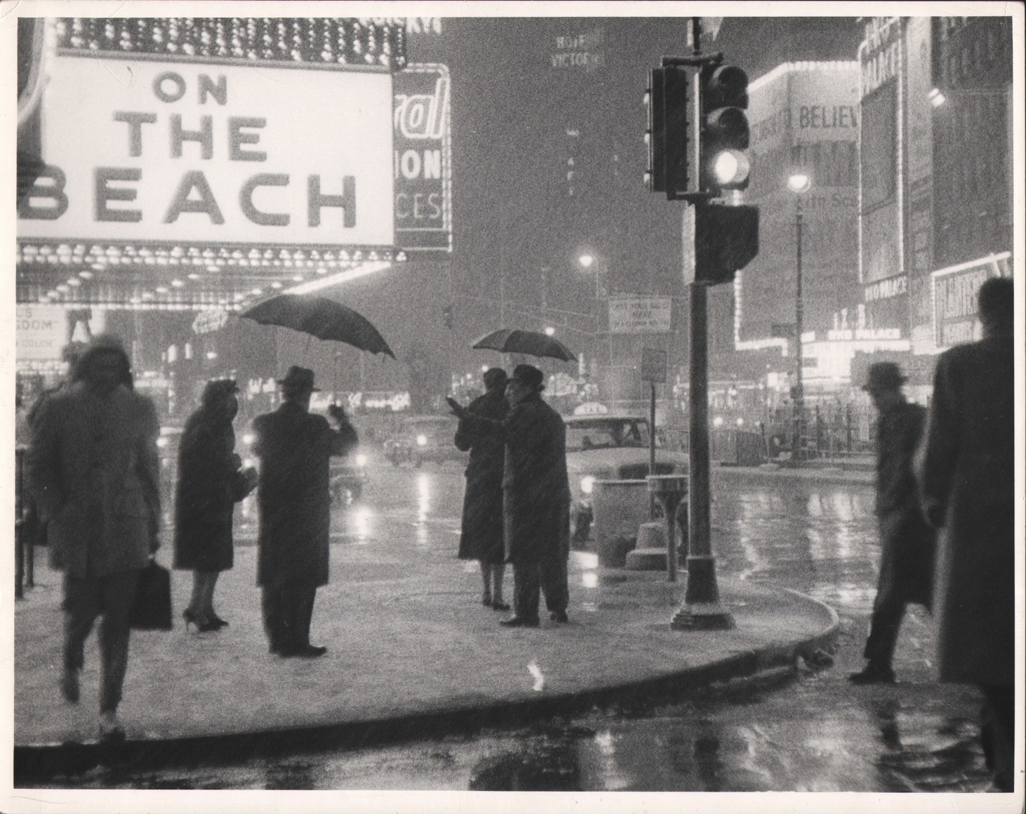 14. Bedrich Grunzweig, Times Square at Night, New York City, ​c. 1959. Pedestrians stand in the sidewalk and street holding umbrellas; a large marquee in the upper left of the frame reads &quot;On the beach&quot;.