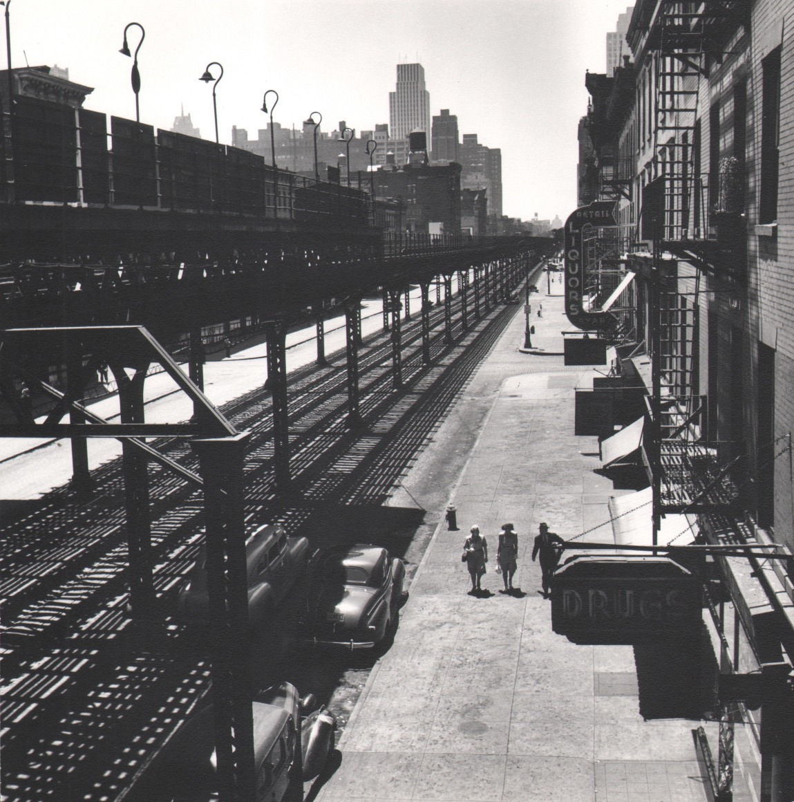 45. Esther Bubley, View of Third Avenue El looking downtown from 53rd Street. The El goes as far downtown as the Battery, ​1946. Elevated view of train tracks and three pedestrians walking on the sidewalk on the right of the frame.
