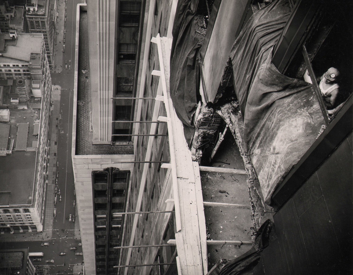 04. Anonymous, Beginning Repairs on Empire State, ​1945. Above view of mid-repair building facade with rebar, tarp, and wood planks visible. A construction worker can be seen on the far right.