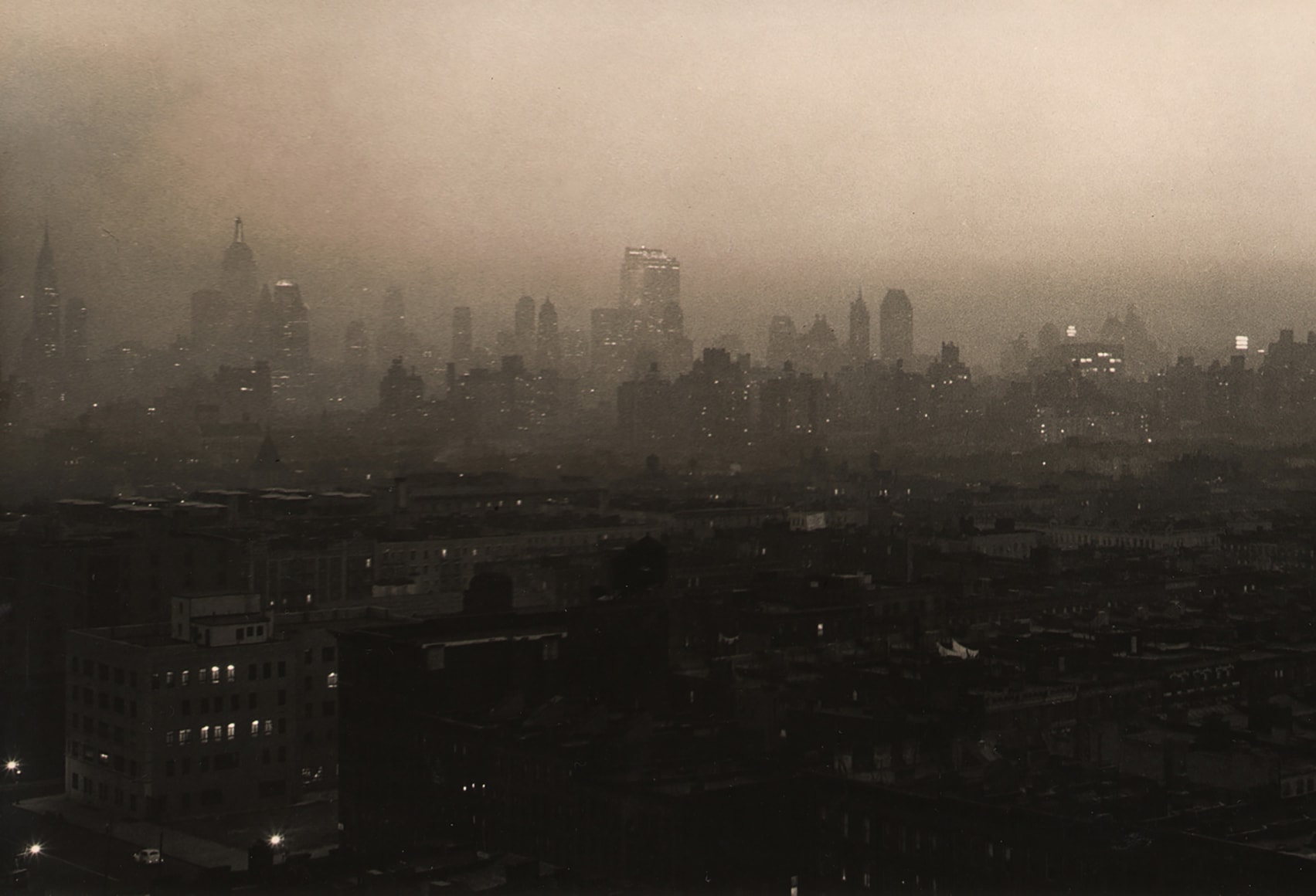 Paul J. Woolf, New York Skyline Evening Haze, ​c. 1936. Dark and hazy cityscape with skyscrapers in the distance silhouetted across the frame.