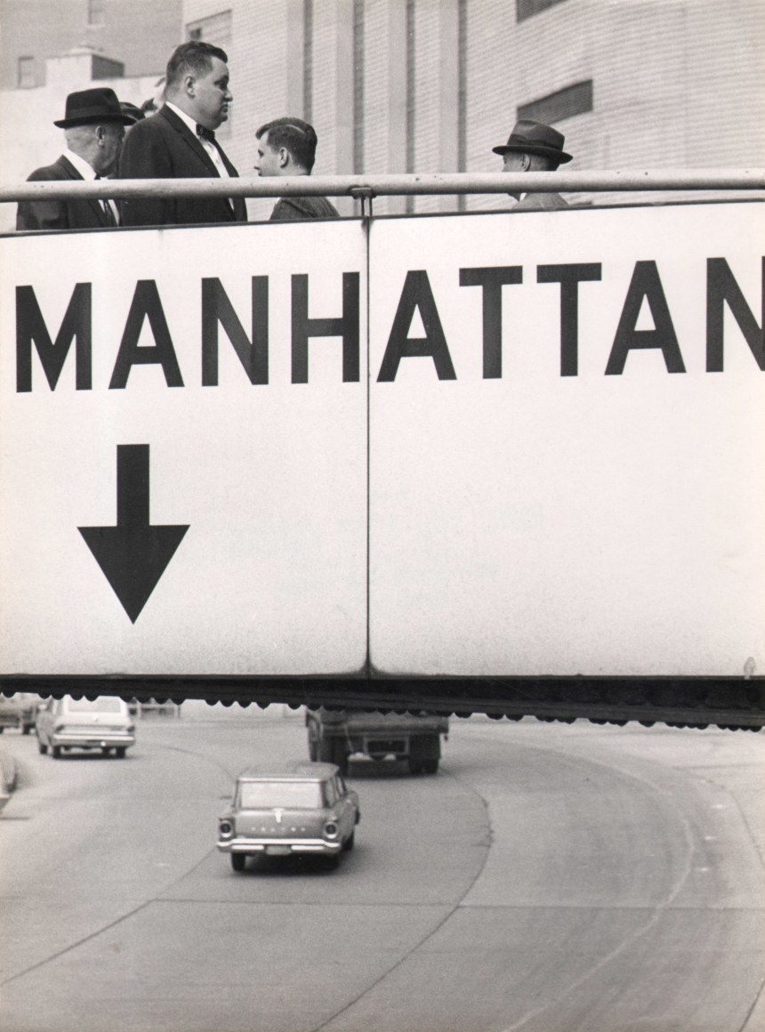 09. Jan Lukas, Untitled, ​1964. Pedestrians walk over a footbridge labeled &quot;Manhattan&quot; above a multi-lane road with cars.
