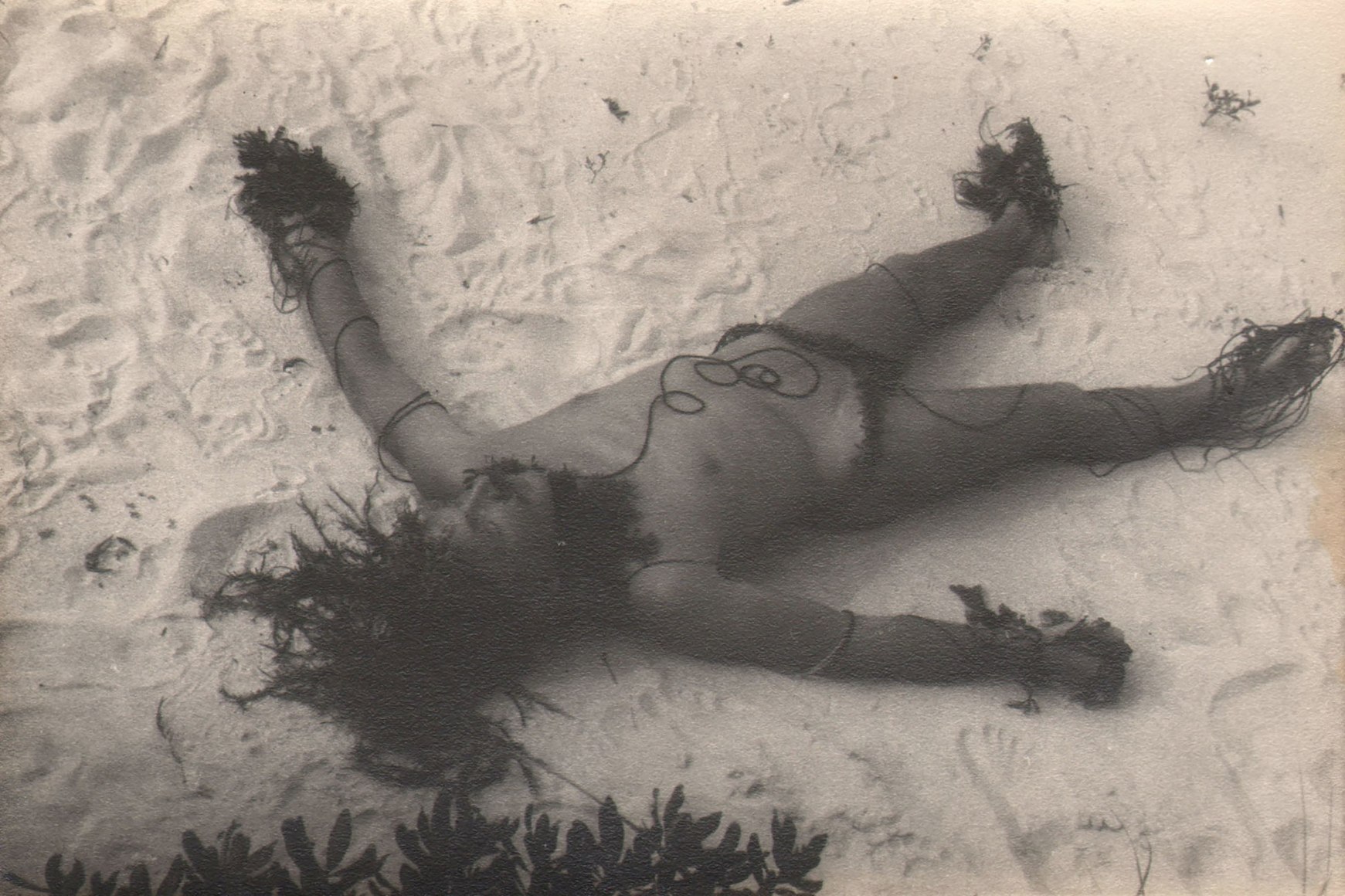 PaJaMa, Jared French, Fire Island, ​1942. A nude male lays in the sand with head in the lower left and legs in the upper right of the frame, seaweed covering his hands, feet, head, neck, and crotch.