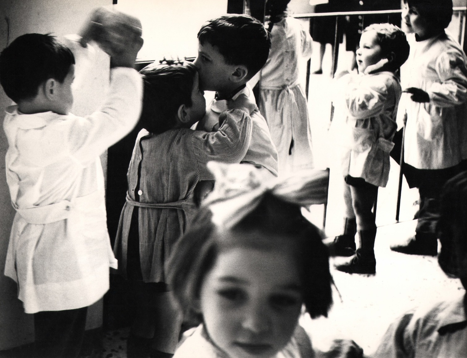 12. Renzo Tortelli, Piccolo Mondo, 1958&ndash;1959. High contrast image. A crowd of children in white gathered in a room. Central boy appears to kiss a smaller boy on the forehead.